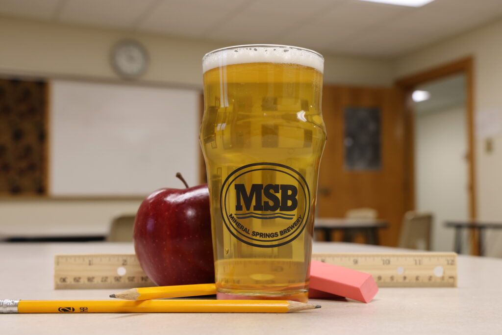 The Dot IPA in front of a whiteboard in a classroom surrounded by pencils, erasers, a ruler, and an apple. All the essential classroom supplies.